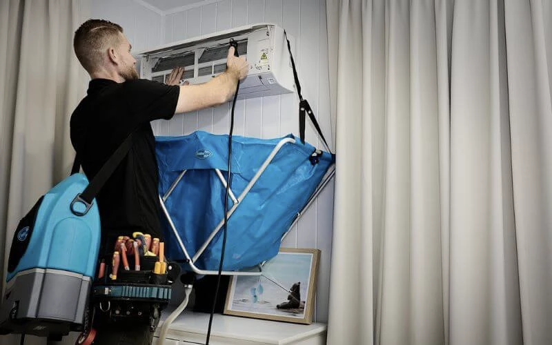 A man with a backpack and a blue bag is working on an air conditioner at home.